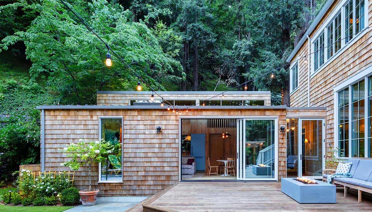 Cedar shingle guest cottage at dusk with open sliding doors, string lights, fire pit, and deck seating