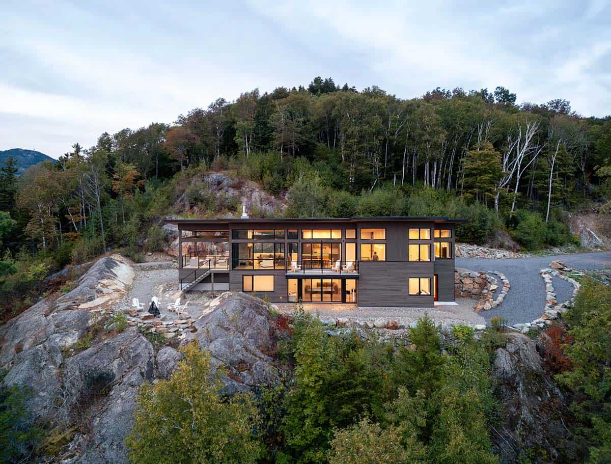 Aerial dusk view of illuminated two-story modern home perched on rocky hillside surrounded by forest
