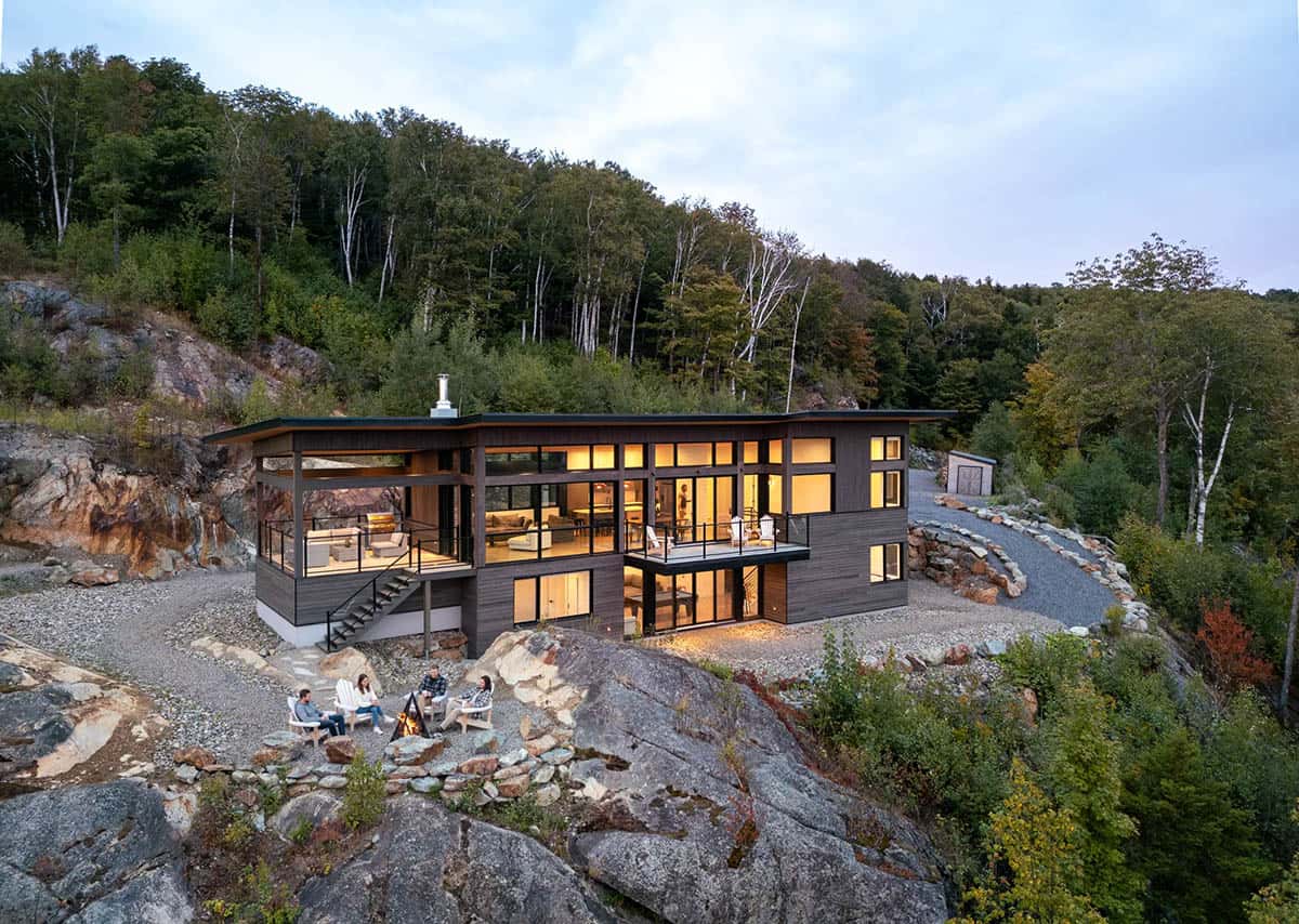 Family gathered around campfire on rock ledge in front of illuminated modern home at dusk