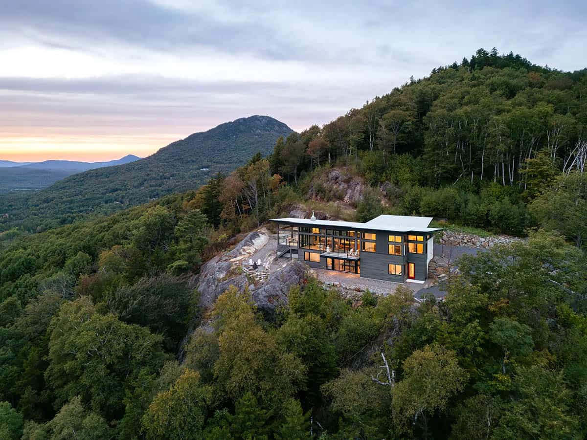 High aerial view of lit modern home on rocky hilltop with mountain range at sunset