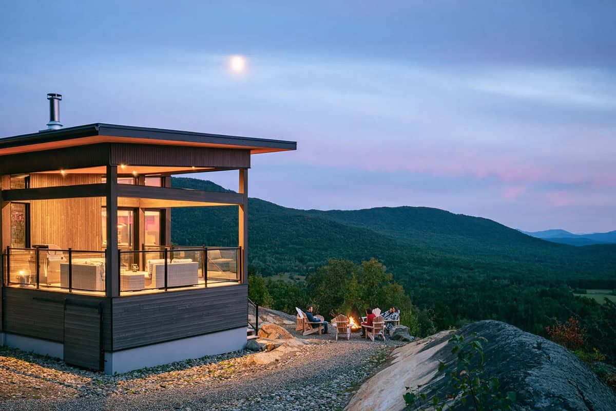 Modern cliffside house glowing at twilight with campfire gathering on rocky ledge and mountain silhouette backdrop