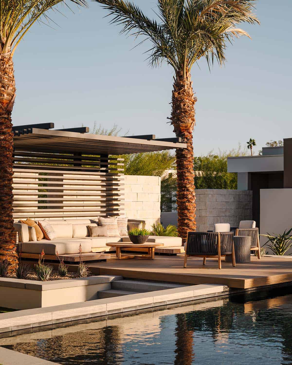 Outdoor lounge area with cream sectional sofa, teak coffee table, and modern pergola beside a rectangular pool, framed by tall palm trees