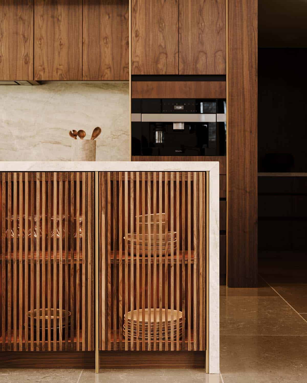 Close-up of kitchen island with slatted wood cabinet fronts, brass trim, and white stone countertop with wall-mounted Miele appliances behind
