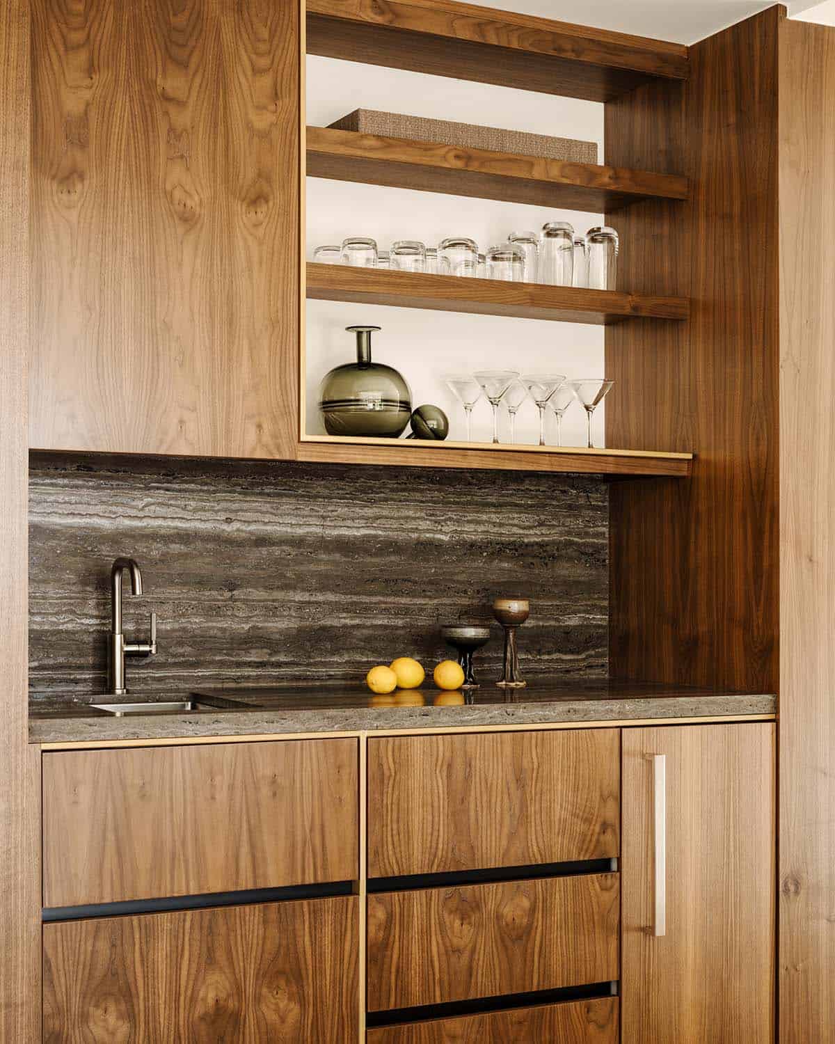  Wet bar niche with walnut cabinets, dark travertine backsplash, open shelving with glassware, and brass accents