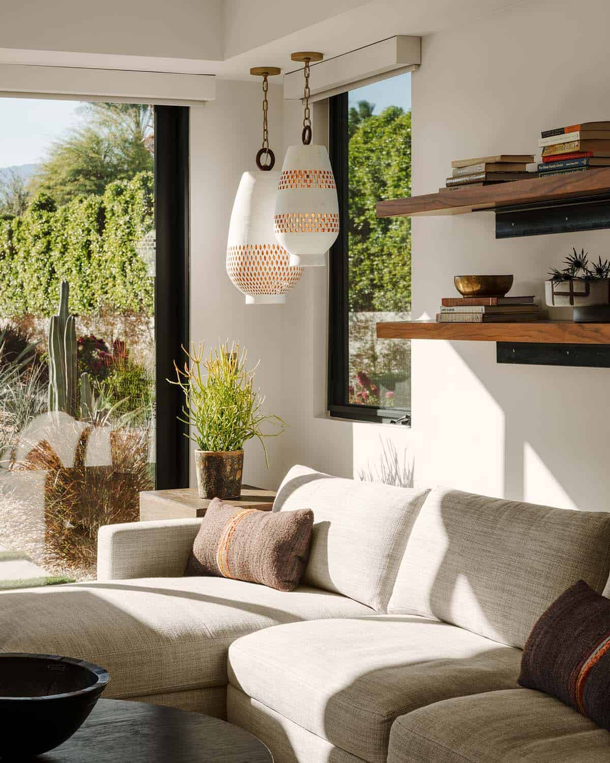 Bright sitting room corner with white ceramic pendant lights, floating walnut shelves, cream sofa, and desert garden view
