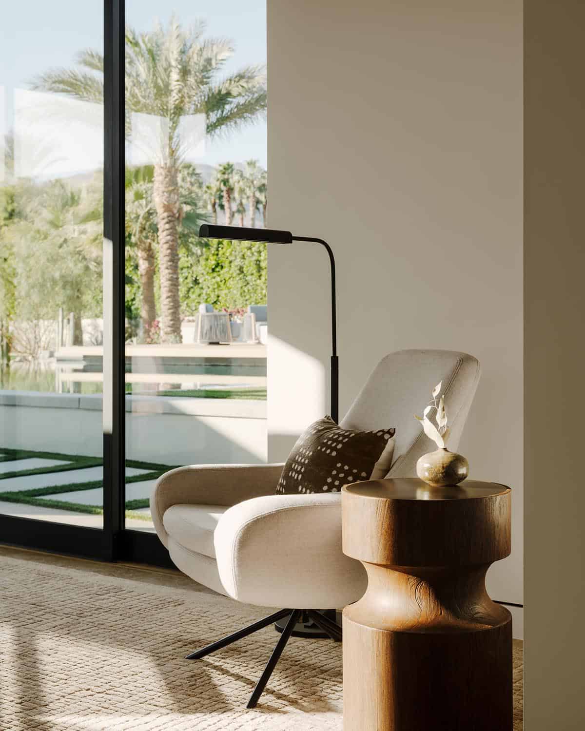 Reading nook with curved cream accent chair, sculptural stacked wood side table, black floor lamp, and floor-to-ceiling pool view