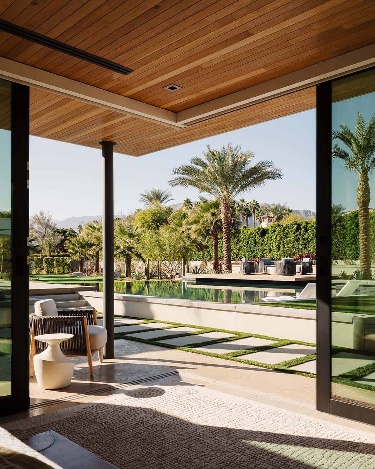 Covered terrace with wood-plank ceiling opening to a long reflecting pool, putting green, and mountain backdrop lined with palm trees