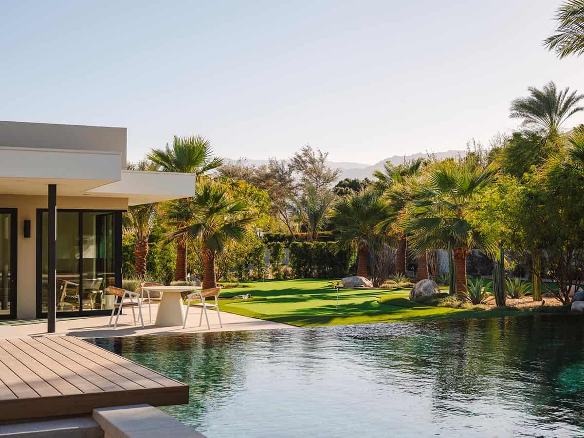 Backyard view from pool deck showing flat-roof modern home, putting green, desert landscaping, and mountain silhouette at dusk