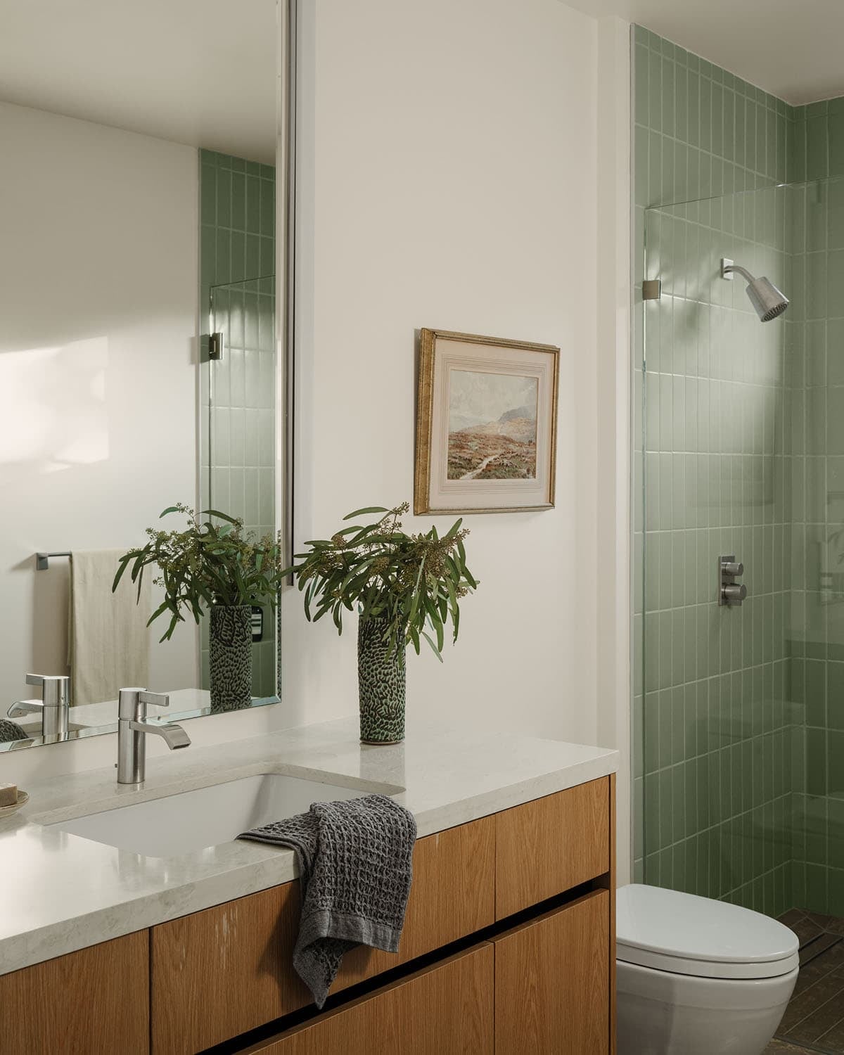 Guest bathroom with white quartz vanity, oak cabinet base, sage green vertical tile shower, and framed landscape artwork