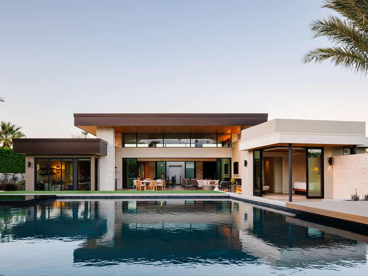 Rear exterior of modern desert home at dusk with flat rooflines, stone and stucco cladding, and long reflecting pool in foreground