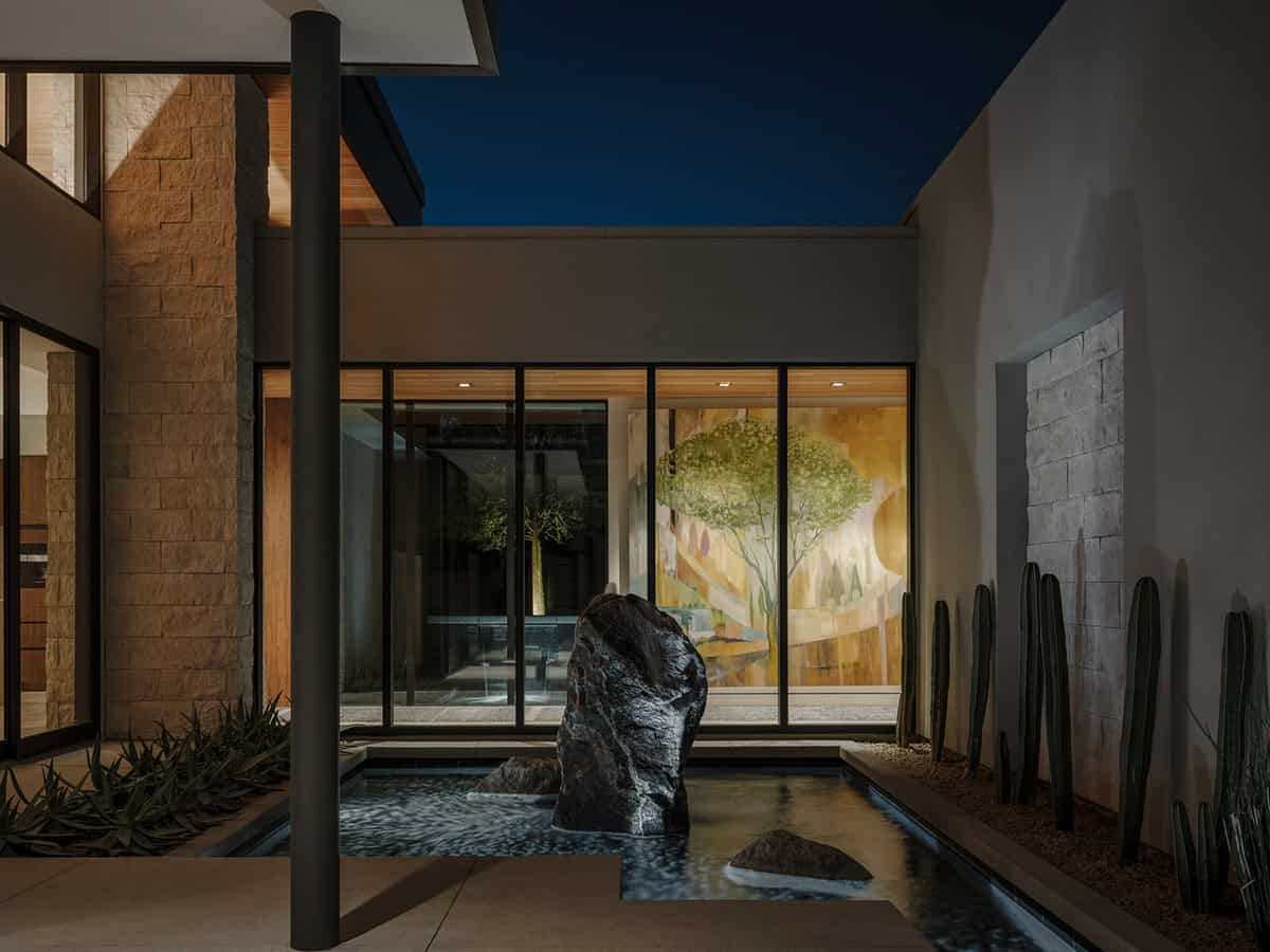 Courtyard at night with a boulder water feature, agave, cacti, and a landscape painting visible through glass walls