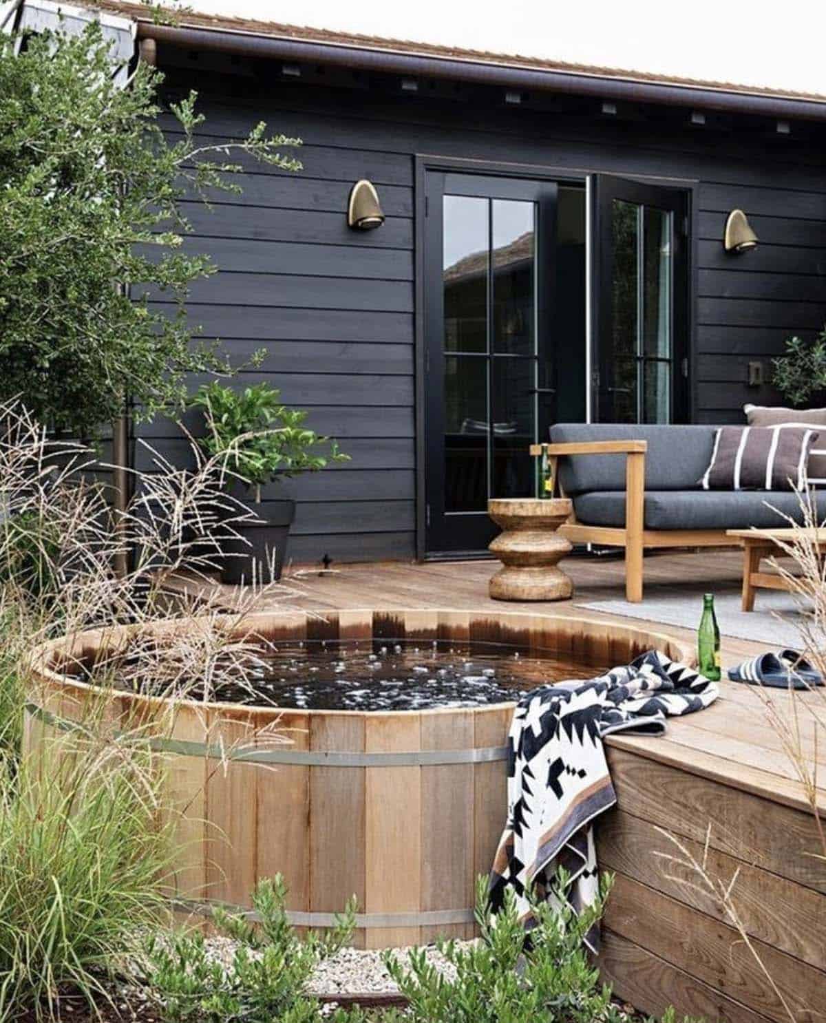 A wooden barrel hot tub deck beside a modern black cabin with a teak lounge chair and ornamental grasses