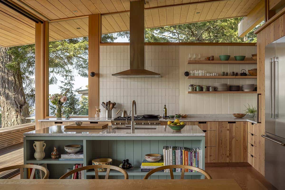 Open kitchen with sage green island, open shelving, white tile backsplash, and forest views through wraparound windows