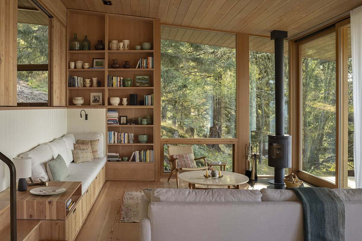 Living room with built-in wood bookshelf, window seat, wood stove, and forest views through corner windows