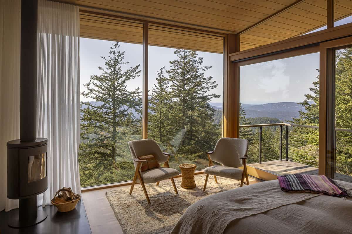 Primary bedroom with wood stove, bouclé lounge chairs, and floor-to-ceiling windows overlooking forested hillside