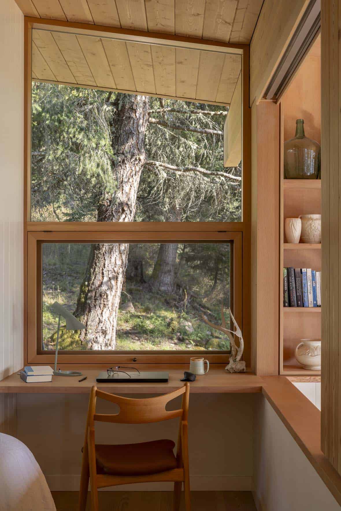 Built-in wood desk nook with large forest-view windows and open shelving with ceramic vessels