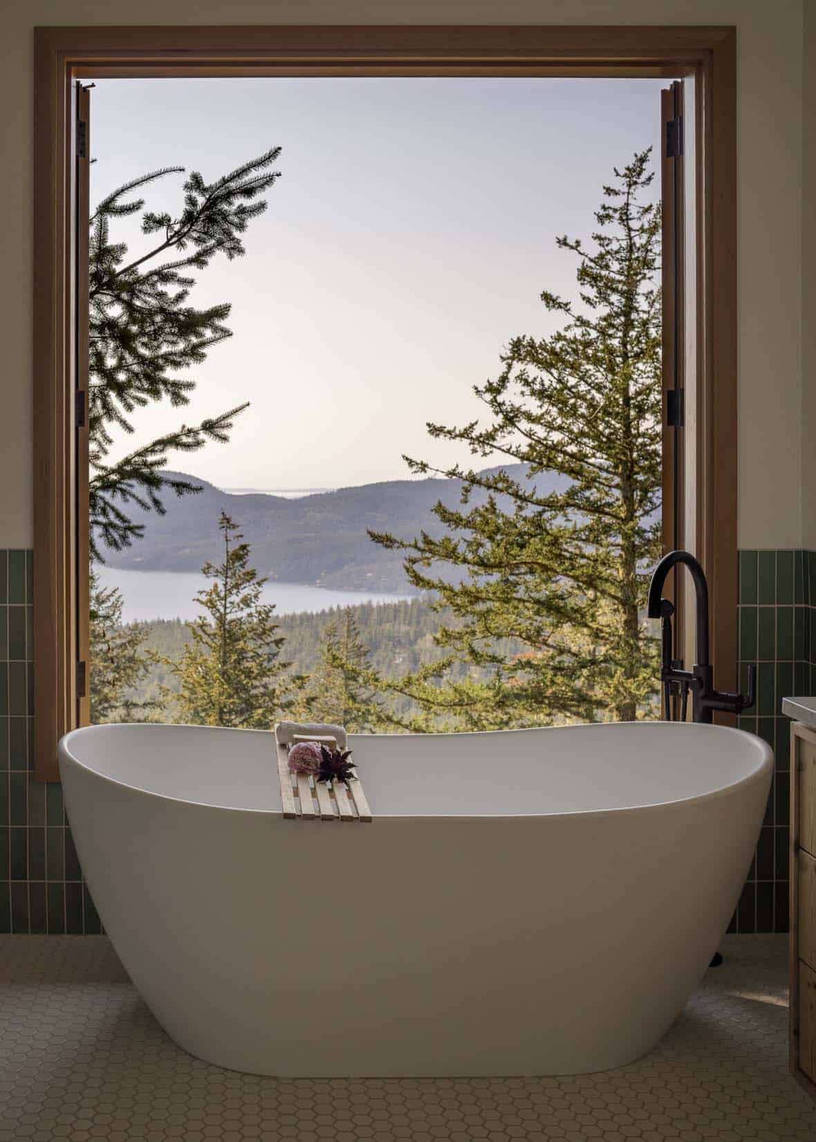 Freestanding soaking tub with floor-to-ceiling window framing treetop and water views, green tile walls