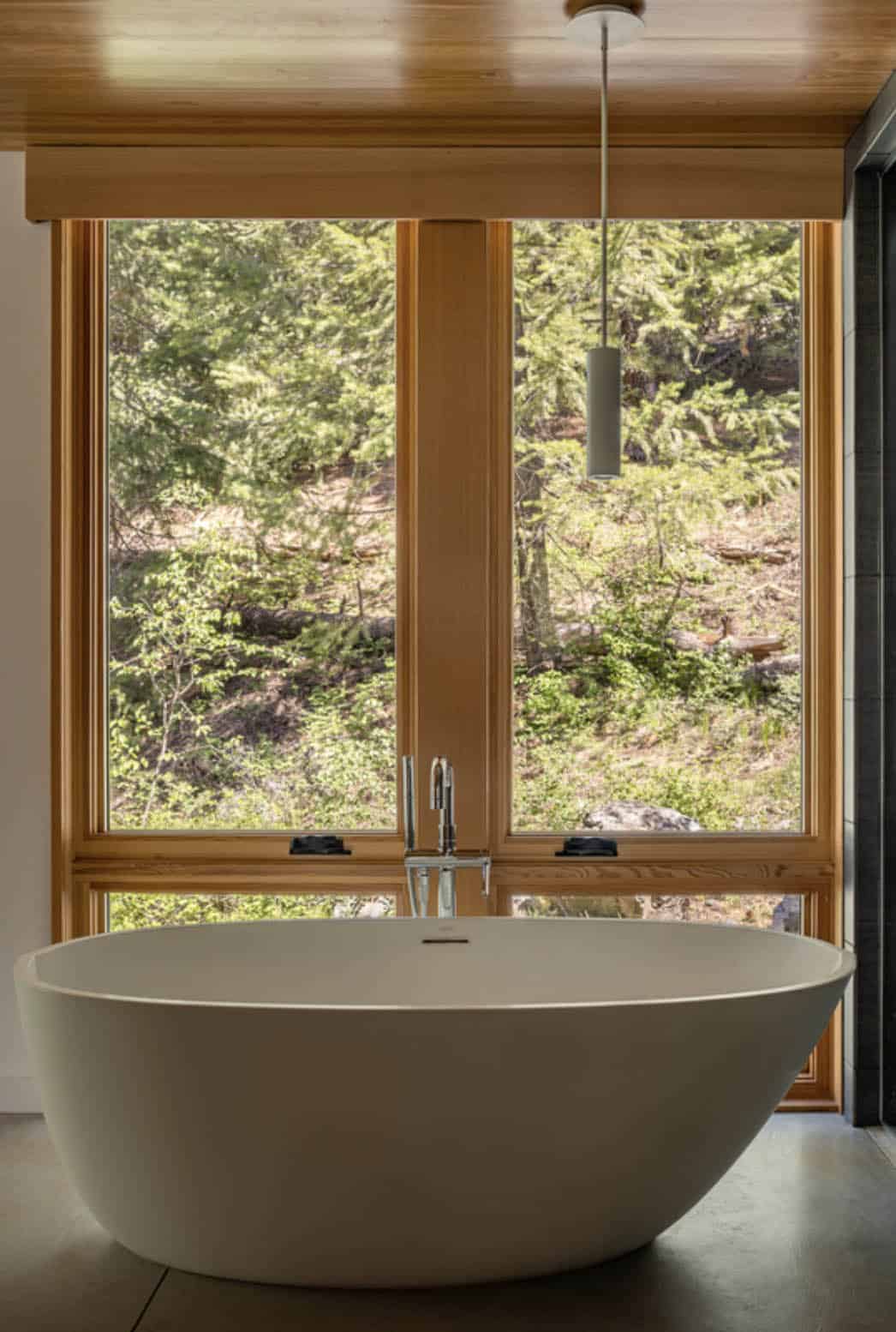 Freestanding soaking tub in front of large wood-framed windows overlooking forest
