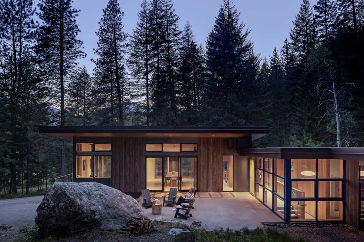 Twilight exterior of modern hillside house with glowing interior, boulder patio, and person seated by fire