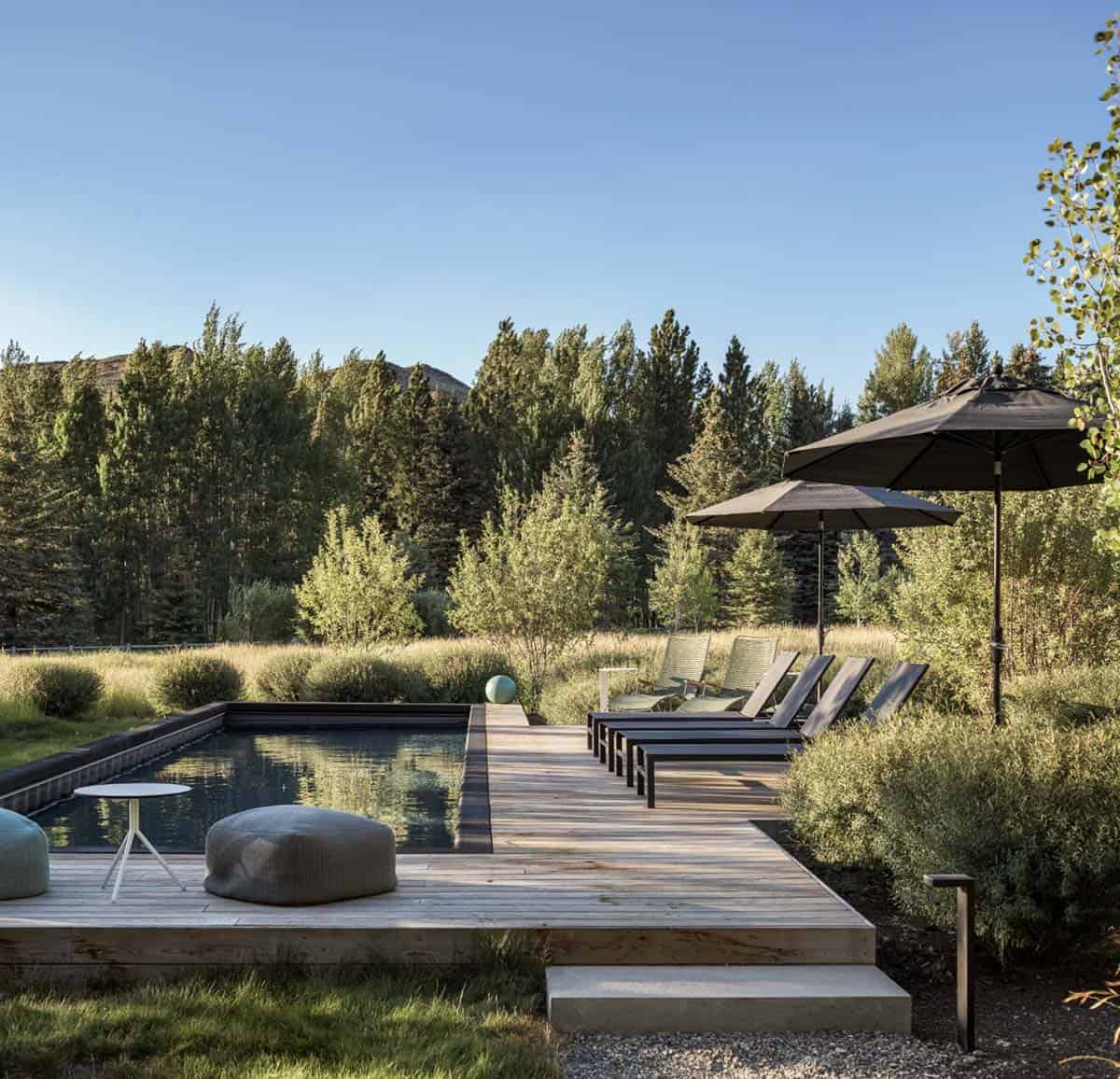 Wooden pool deck with dark lounge chairs, umbrellas, and a meadow backdrop