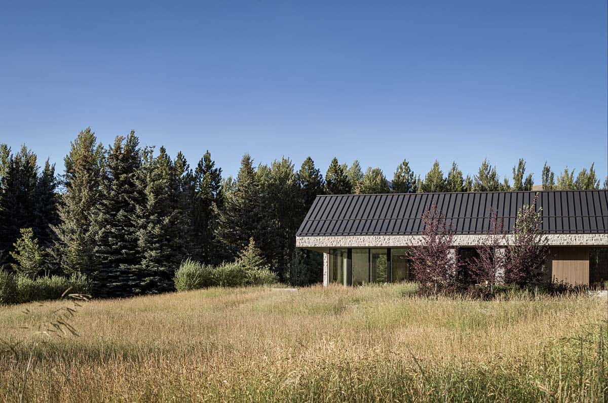 A stone and timber house with a dark metal gable roof seen across a tall grass meadow