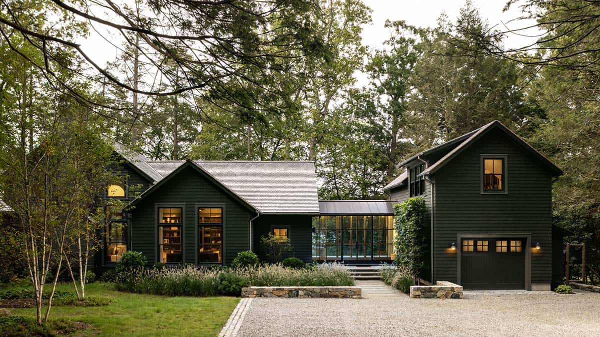 Dark green shingle-style lake house exterior with attached garage and gravel driveway, surrounded by trees