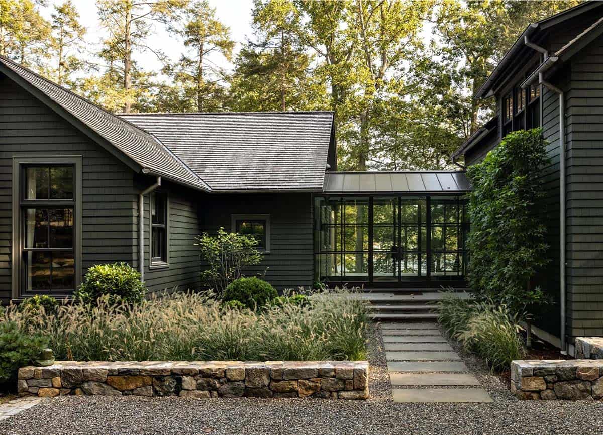 Side view of dark green lake house showing glass-walled entryway and stone retaining wall garden beds