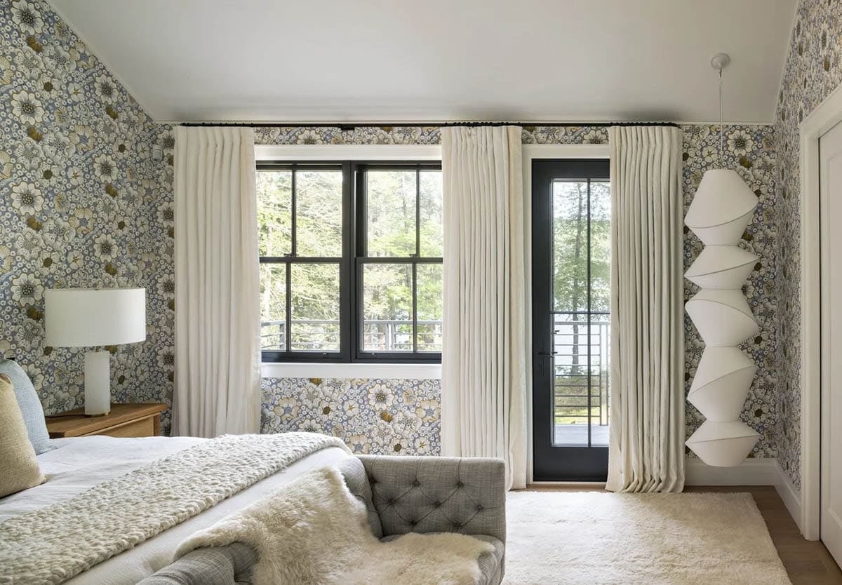 Bedroom with blue floral wallpaper, cream curtains, and black-framed door to balcony