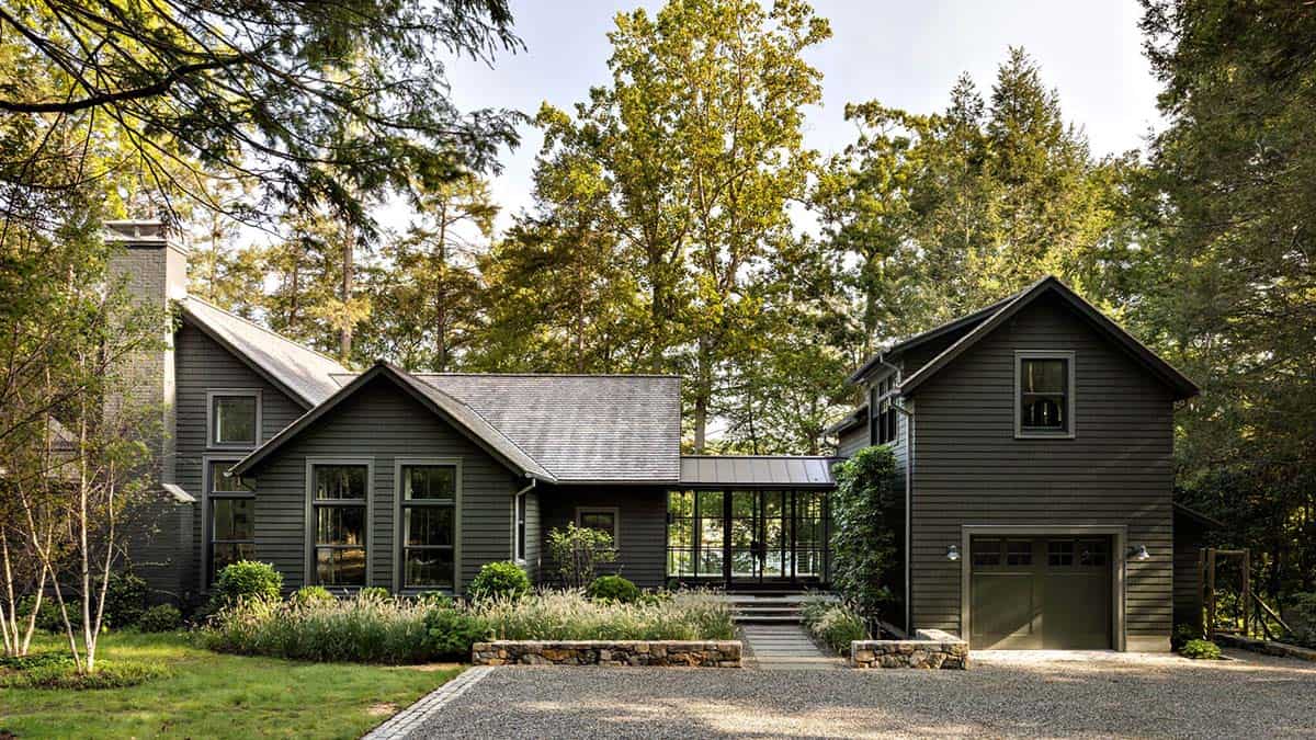 Front view of dark green lake house with stone chimney, garage, and gravel driveway