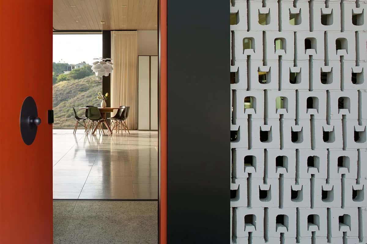 View through open orange front door revealing dining area with Eames chairs and floor-to-ceiling hillside views