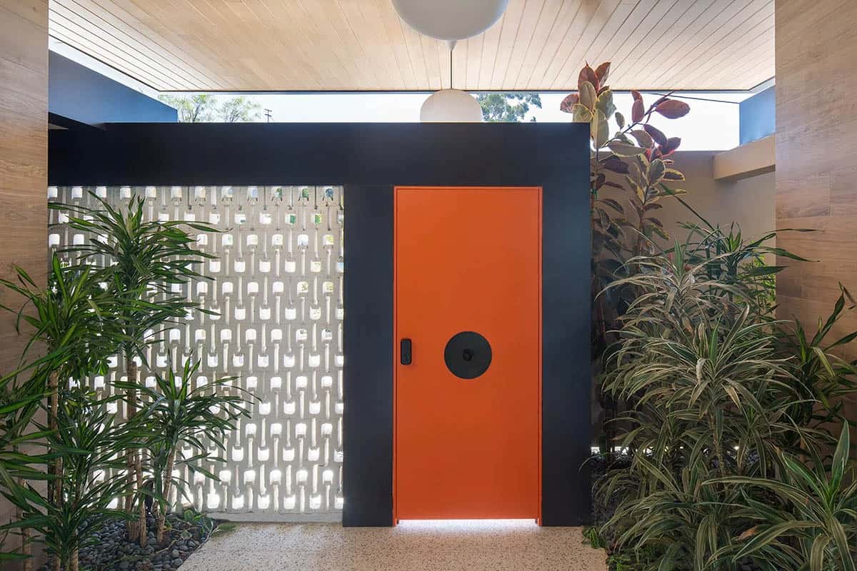 Close-up of orange pivot front door framed in dark steel with globe pendant light and flanking tropical plants