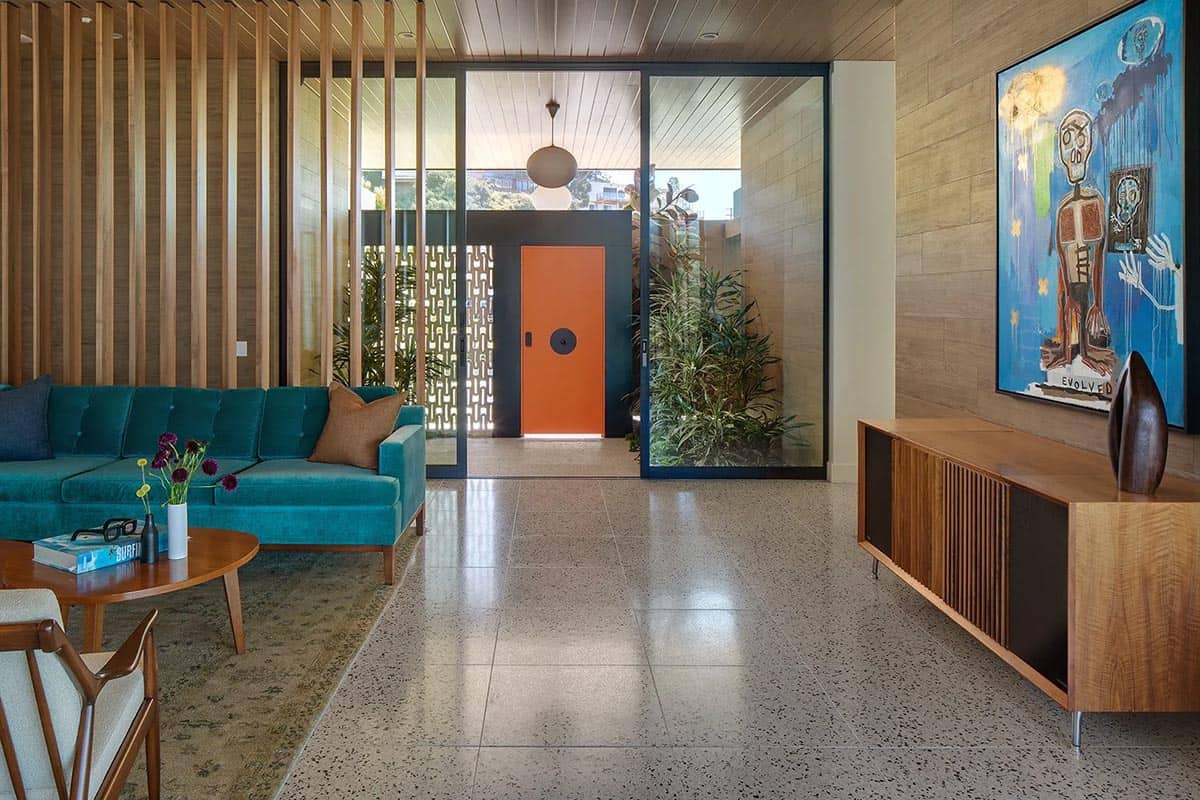 Living room looking toward entry with teal velvet sofa, wood slat wall, terrazzo floors, and large-scale wall art