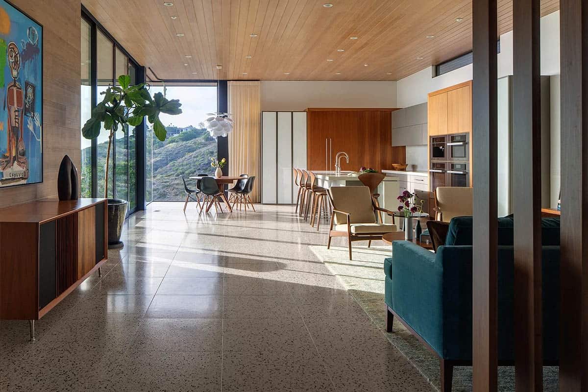 Open-plan living, dining, and kitchen with wood ceiling, terrazzo floors, walnut cabinetry, and panoramic hillside views