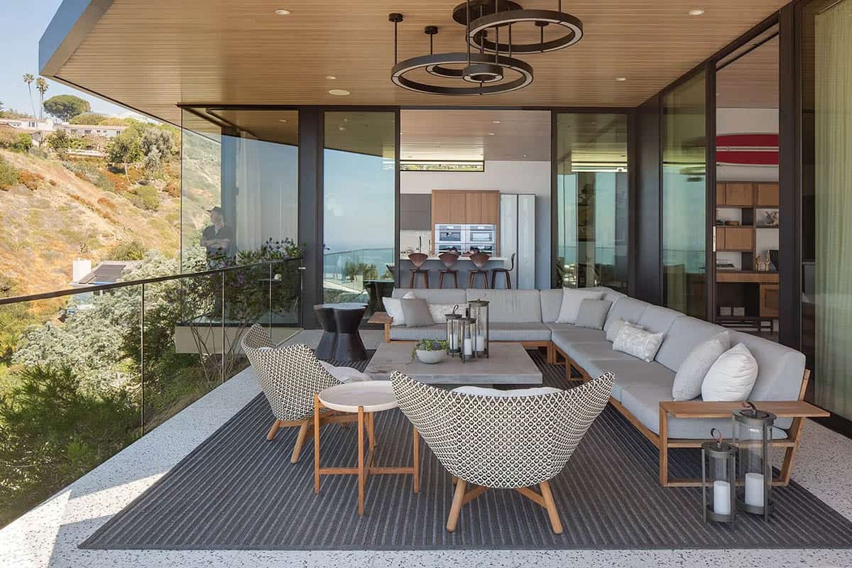 Covered terrace with sectional seating, woven accent chairs, and glass railing framing canyon and ocean views