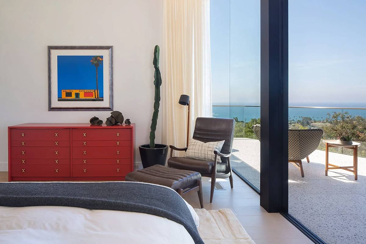 Primary bedroom with red lacquer dresser, leather lounge chair, cactus plant, and sliding glass door to ocean-view balcony