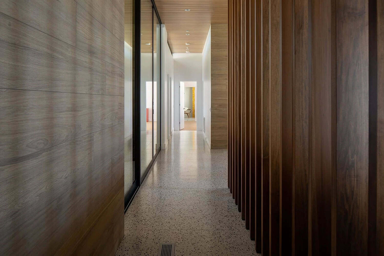 Hallway with wood slat partition, large-format wood-look wall tiles, terrazzo floors, and glimpse of yellow-tiled bathroom