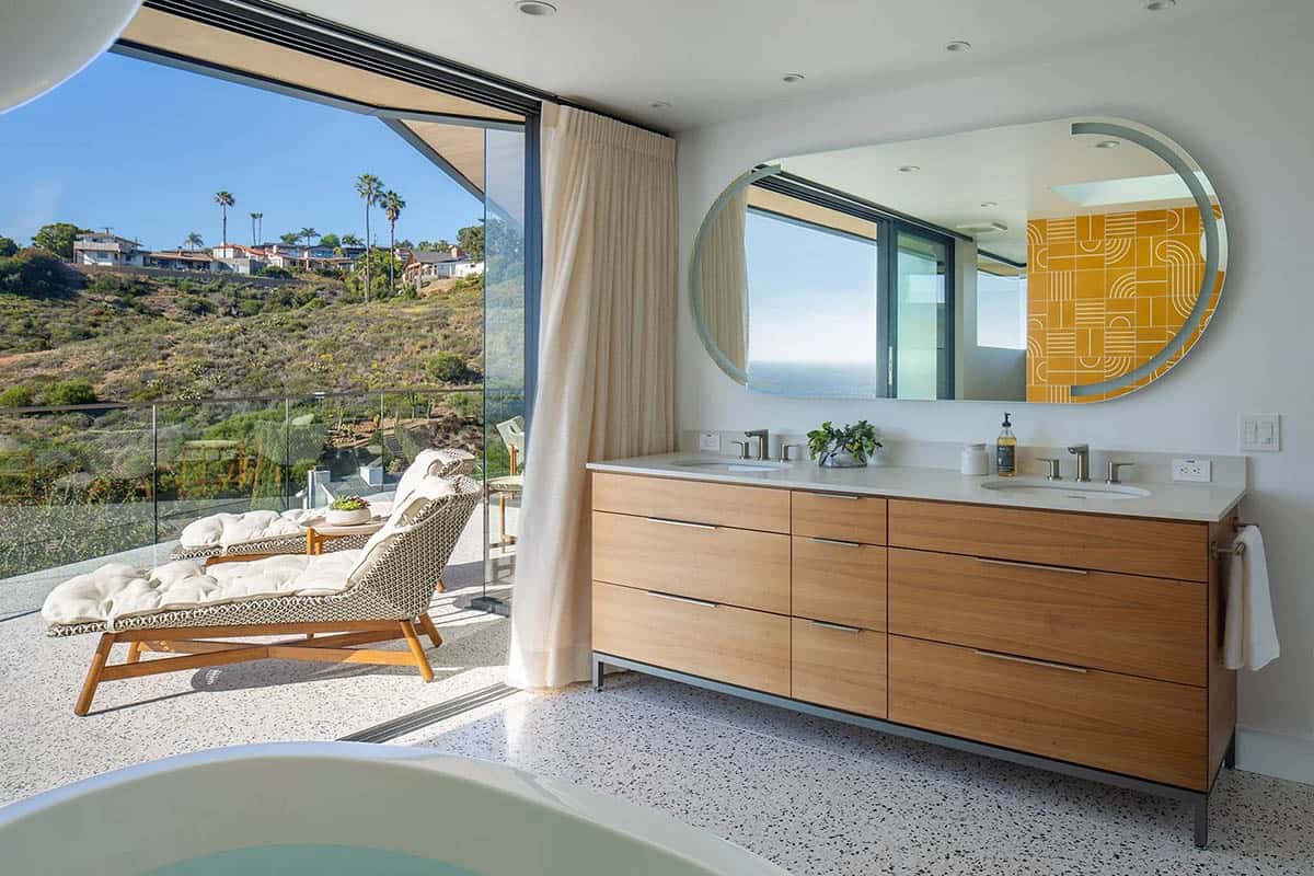 Primary bathroom double vanity with wood cabinetry, round mirror, and hillside and ocean views through full-height glazing