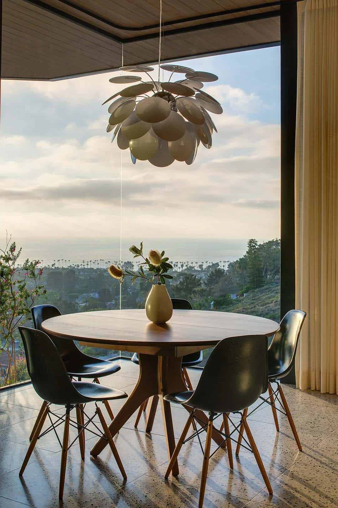 Dining area with round walnut table, Eames shell chairs, sculptural pendant light, and sweeping Pacific Ocean view at dusk