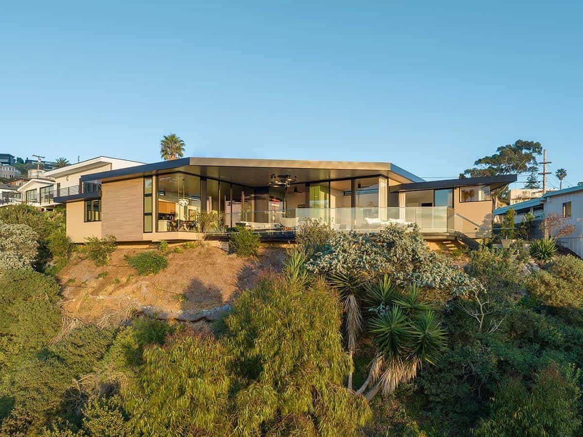 Rear aerial view of flat-roofed mid-century home perched on hillside with glass walls, covered deck, and native vegetation