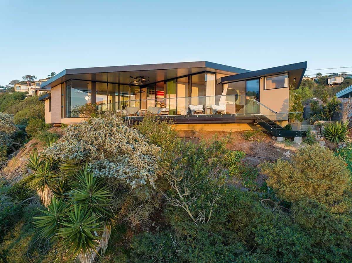 Angled rear exterior view of mid-century modern hillside home with wraparound floor-to-ceiling glass walls, glass-railed deck, and native coastal scrub in the foreground