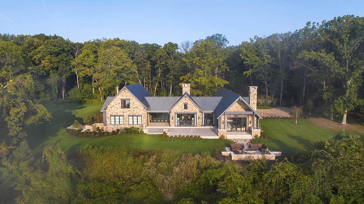 Aerial view of rear facade with stone gables, dark standing-seam roof, and patio overlooking wooded property