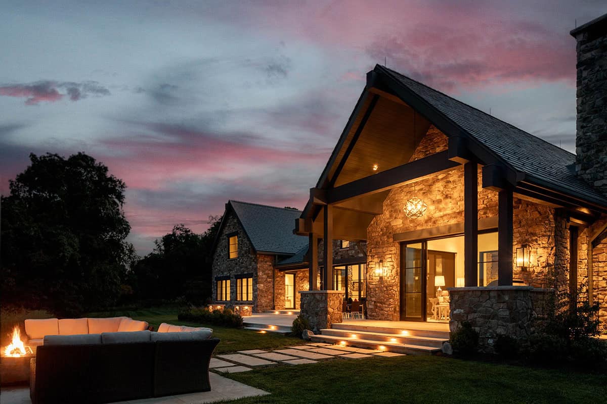 Stone and timber mountain home glowing at dusk under a pink sunset sky with lit fire pit in foreground