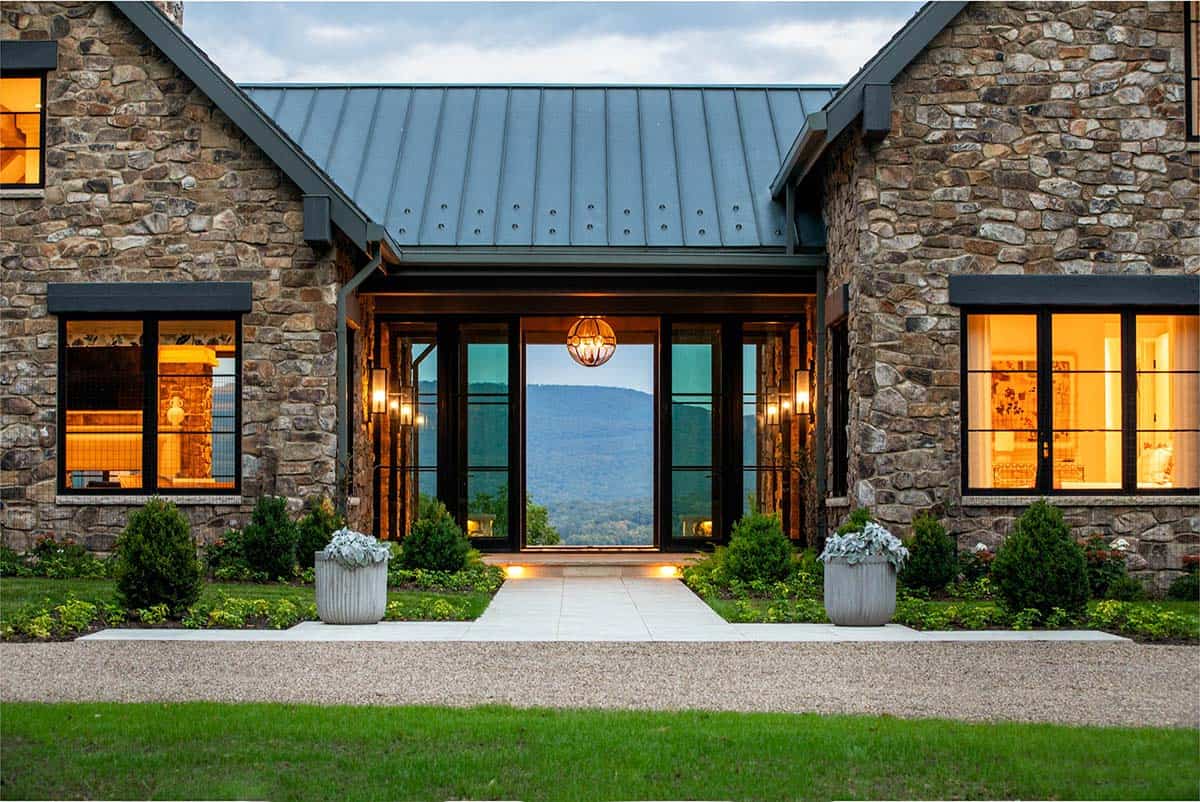 Stone farmhouse-style entry with standing-seam metal roof and glass doors framing a mountain view at dusk