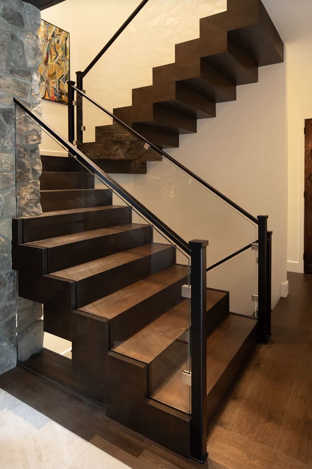 Modern floating wood staircase with black metal and glass railing against a stone wall