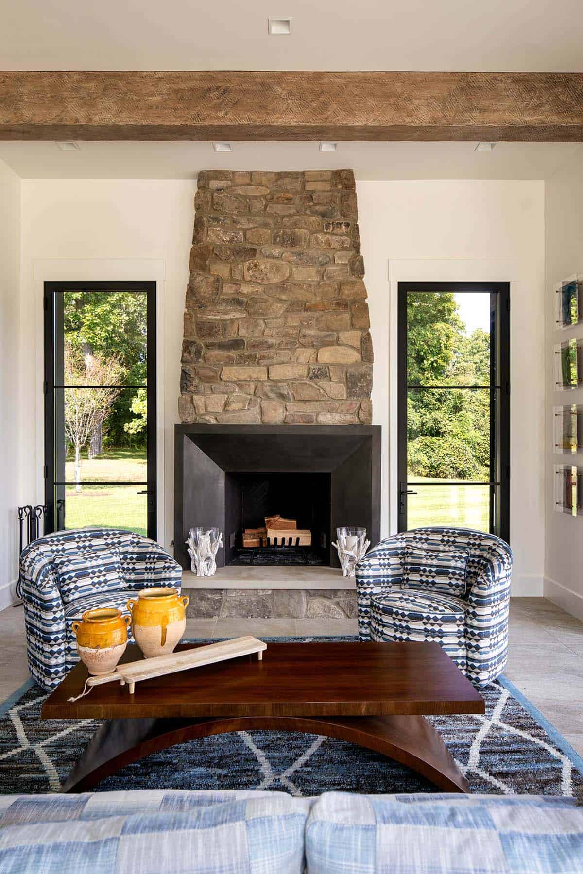 Living room with tall stone fireplace, patterned swivel chairs, and wood beam ceiling flanked by glass doors