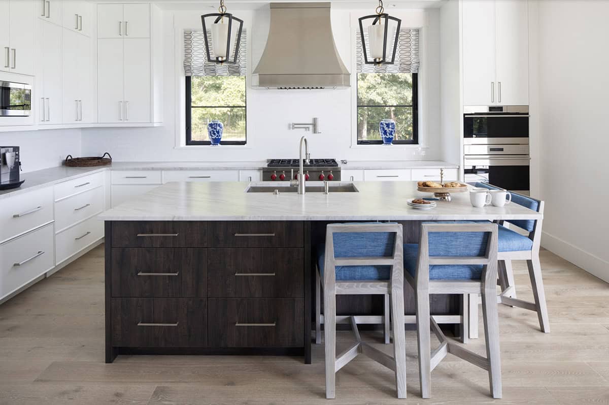 White kitchen with dark wood island, marble countertops, stainless range hood, and blue upholstered counter stools