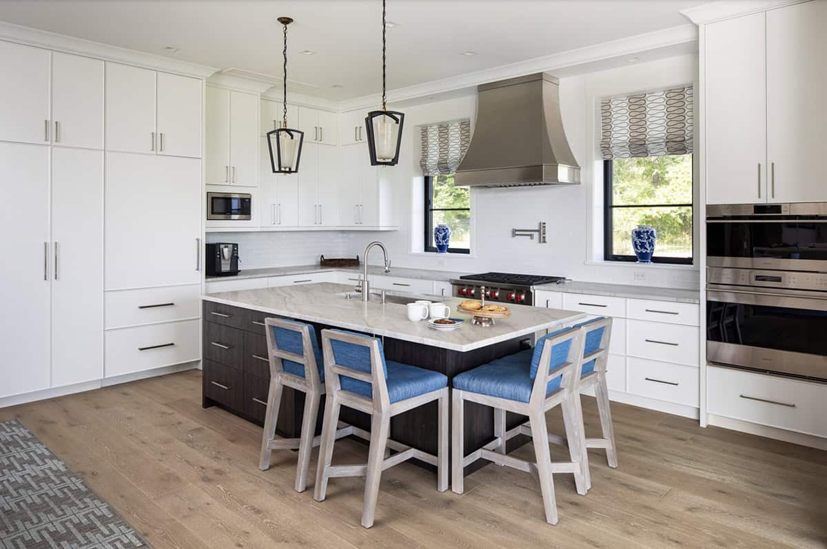 White kitchen with marble-topped island, blue counter stools, and dual wall ovens along stone-accented walls