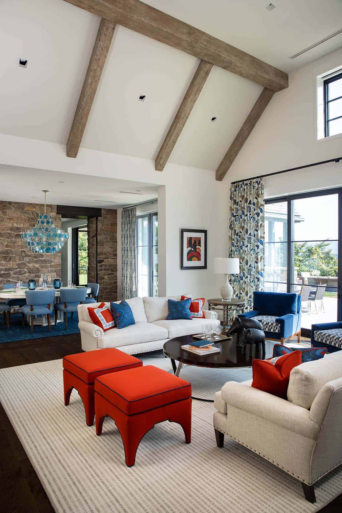 Vaulted living room with wood beams, cream sofas, and orange ottomans opening to a stone-walled dining area