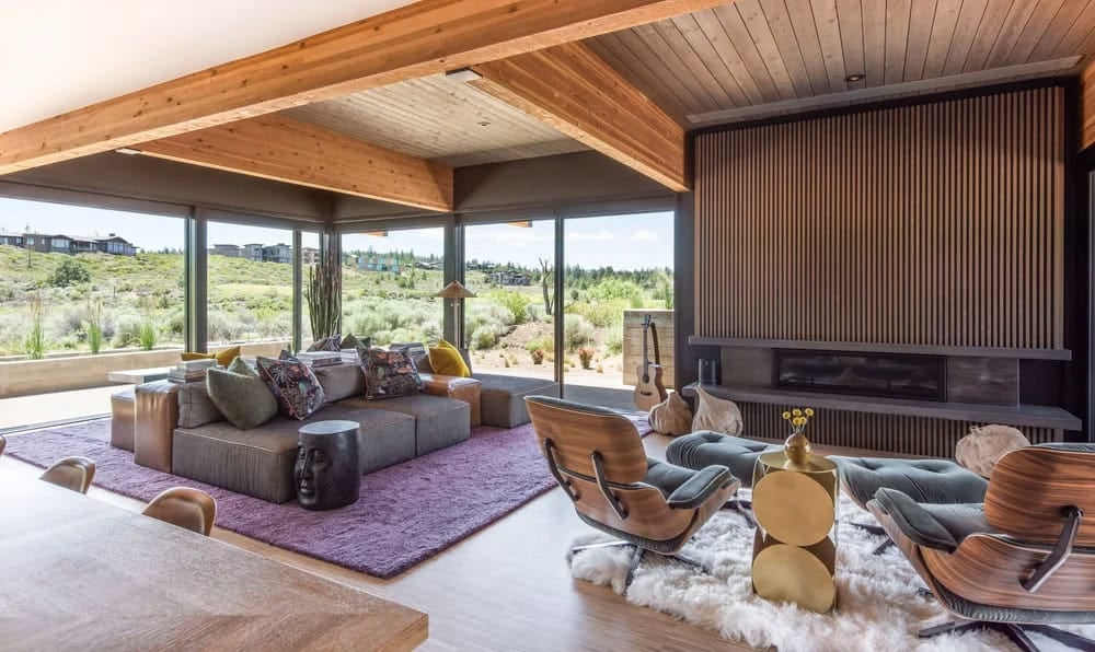 Open-plan living room with wood beam ceiling, floor-to-ceiling windows, gray sectional sofa on purple rug, and vertical wood slat fireplace wall