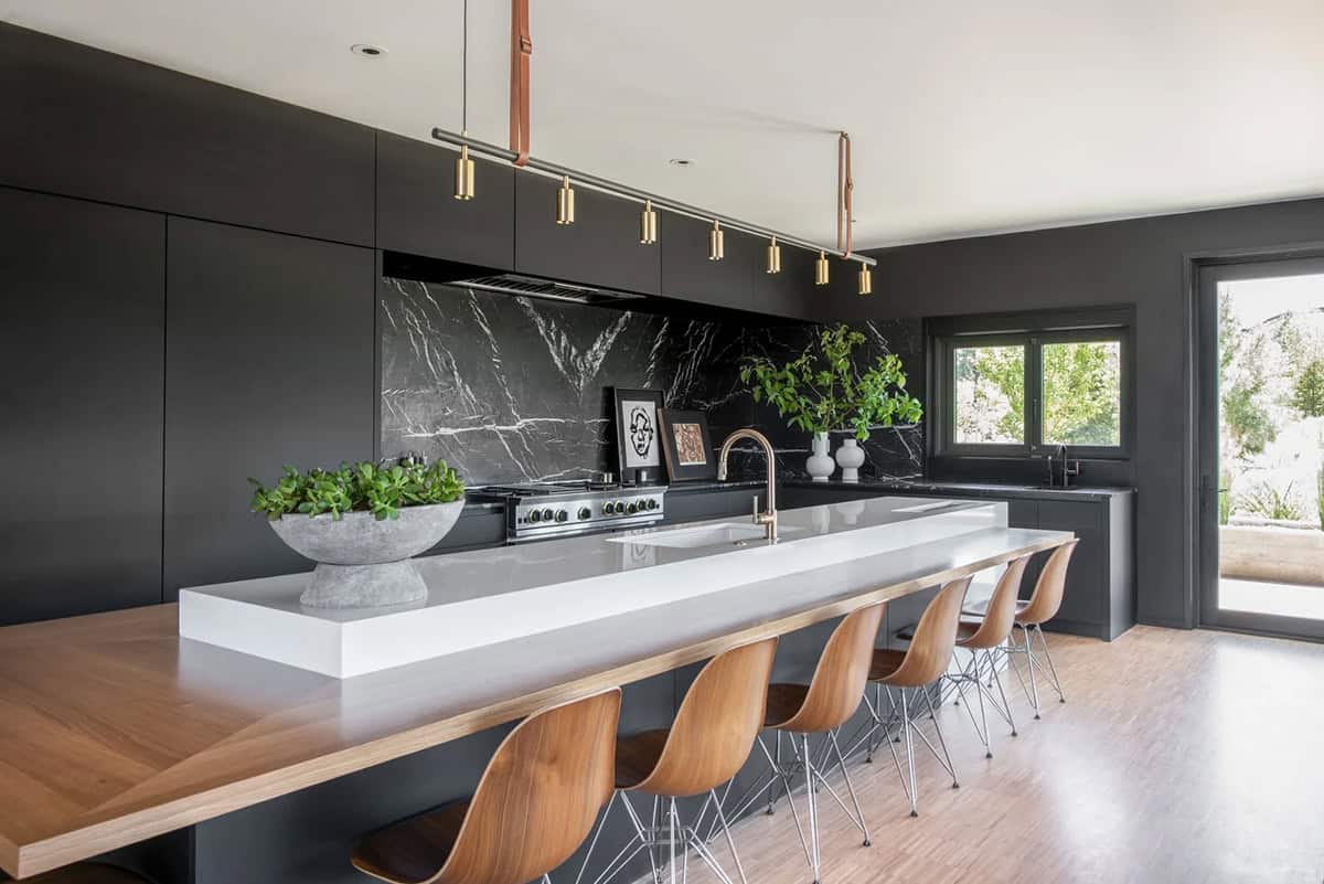 Long white kitchen island with walnut bar stools, brass pendant lights suspended from copper rods, and black marble backsplash
